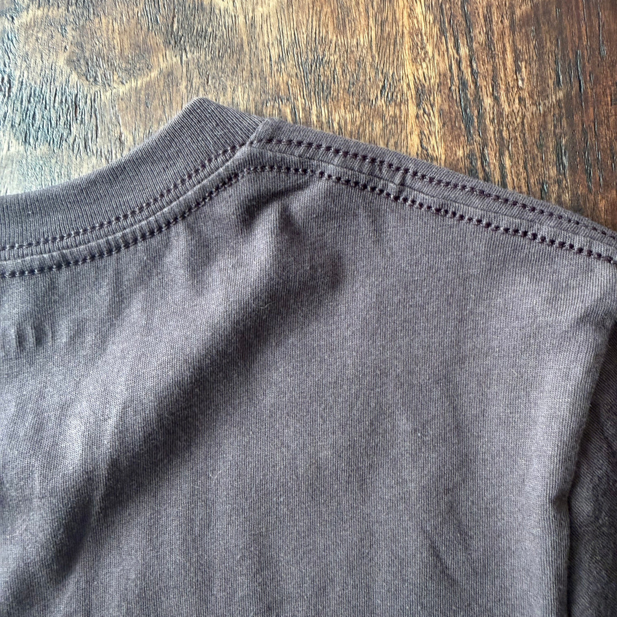 Close-up of a brown crewneck t-shirt laid flat on a rustic wooden surface, showing the fine knit texture and double-stitched collar.

