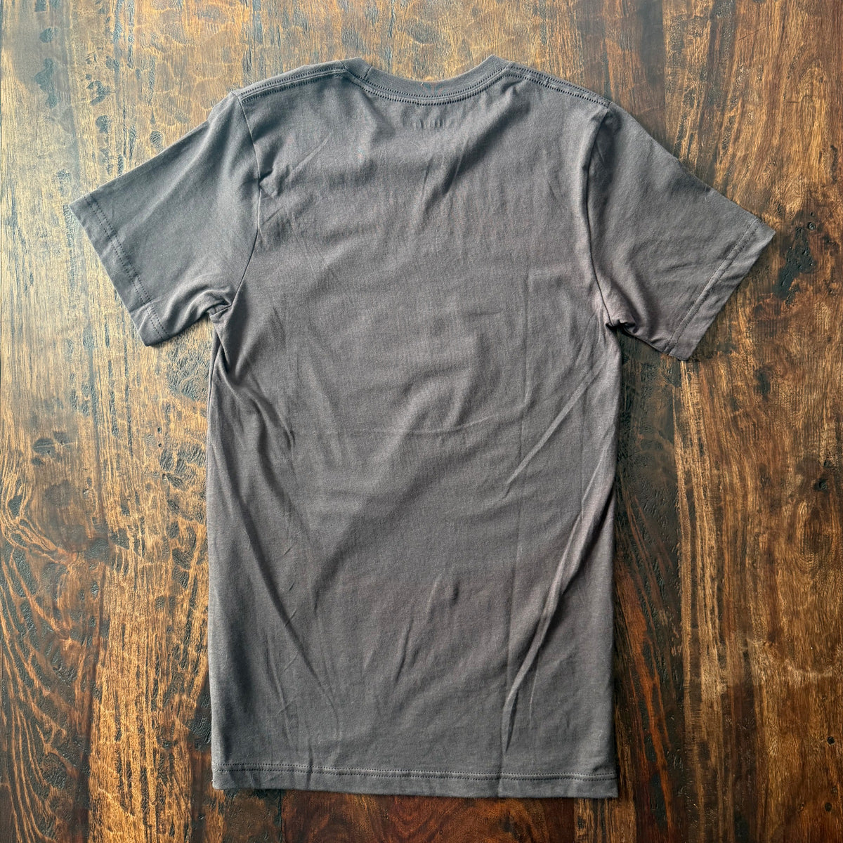 A dark brown short-sleeve t-shirt laid flat on a rustic wooden surface, showing the back side of the garment.
